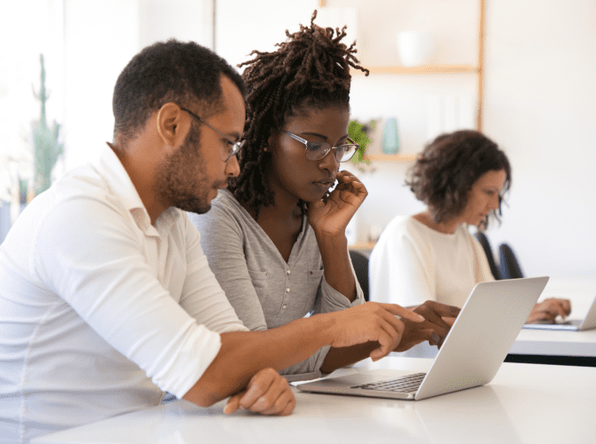 Two people , a male young man and a female young woman watching a laptop. The man is pointing the index finger at the screen,.